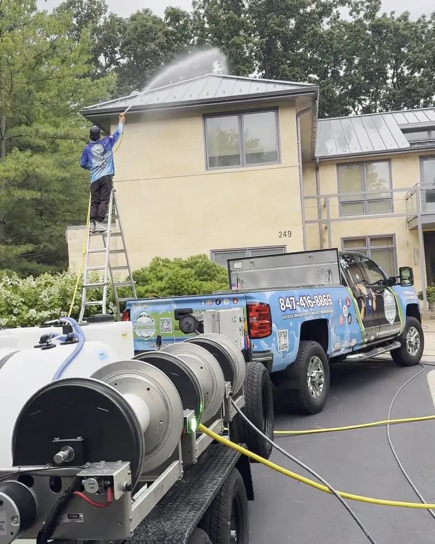 Andrew on a ladder soft washing a roofline, the spray arcing over the gutter, with the Simmons truck in the driveway